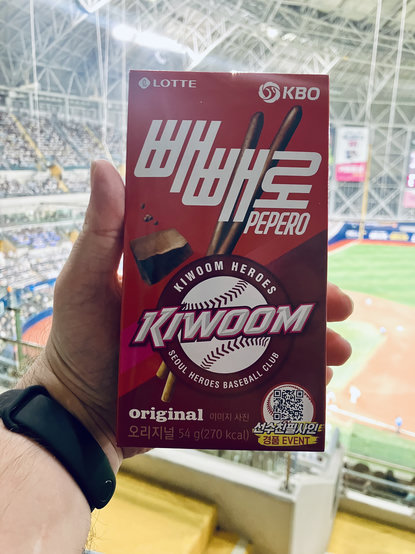 A person holds a special-edition box of Lotte Pepero Original in the foreground while seated inside a baseball stadium. The bright red package features the Kiwoom Heroes logo, KBO branding, and images of chocolate-coated biscuit sticks. A QR code and promotional graphic appear in the lower-right corner of the box. The person’s wrist is wearing a black fitness tracker. In the blurred background, the baseball diamond, players, and grandstands filled with spectators are visible beneath the stadium’s high, steel-trussed roof.