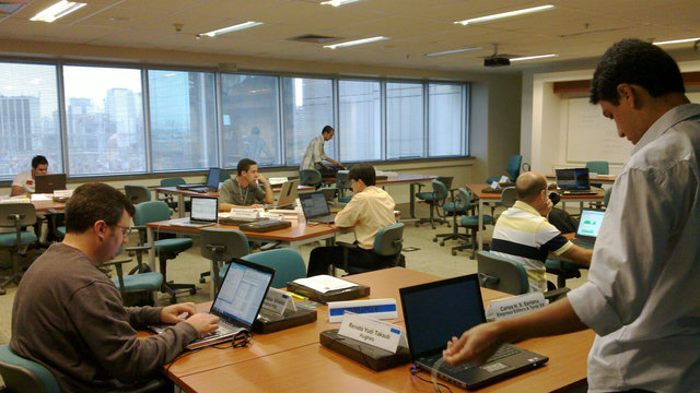 foto mostra uma área de escritórios em um prédio e várias pessoas com laptops em várias mesas