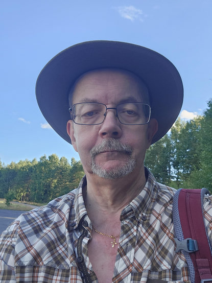 Gubbe i hatt, glasögon, hakskägg och mustach. Rutig skjorta med rem till ryggsäck över vänster axel