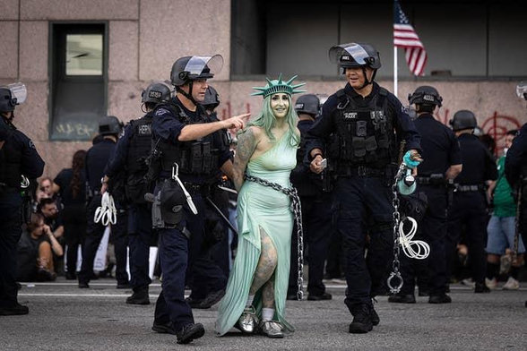 A woman dressed as the Statue of Liberty at a No Kings rally. Police wearing riot gear are putting handcuffs on her.