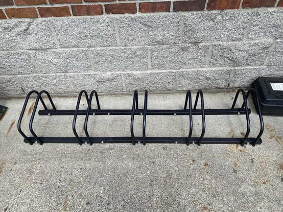 On pavement next to a cement brick and regular-brick building, what looks like a miniature bike rack with five very low-to-the-ground flattened-top pairs of hoops bridging two longer steel(?) bars. It's all painted black. There's a little box off to the right side but it's not related, I think it's some sort of pest control.