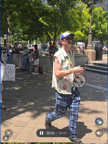 Sam O'Hara, a white man in blue plaid pants, a ballcap, and a white vest covered in anti-Trump pens, walks along a street in DC with other protesters while carrying a bullhorn.