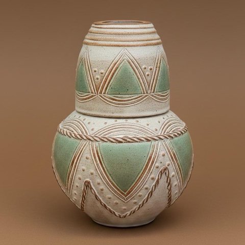 A spherical, decorative ceramic bottle with a small cup on top that acts as a cover when not in use. The piece features a textured off-white surface, aqua accents, and intricate geometric patterns. The background is a plain brown color.