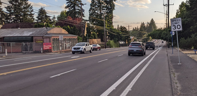 Five lane stroad with buffered and sometimes curb-protected bike lanes, curb-tight sidewalks, sun setting other conifer trees and Mt Tabor.