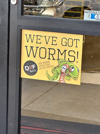A sign on a store door stating "WE'VE GOT WORMS!" with colorful cartoon illustrations of a worm that looks very happy to be about to die, with a fish being carried in its shoulder . With the tagline "OUR WORMS CATCH FISH, OR DIE TRYING