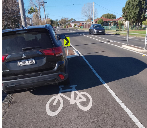 Photo of a white painted bicycle symbol in a parking lane, with a parked car almost on top of it. Immediately in front of the car are lane narrowing concrete islands to force cyclists into the general traffic lane if they're not there already.