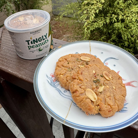 Two peanut butter cookies on a plate. They have a few sichuan peppercorns sprinkled on top. Behind the cookies is a can of Tingly Sichuan Pepper Peanuts from 50hz. If you look closely, you can see there are cookies put into the can, where peanuts used to be. 