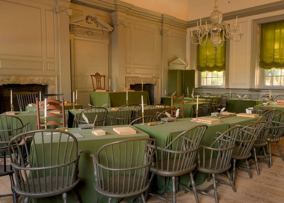 A color landscape photo showing the room inside Independence Hall in Philadelphia where where both the Declaration of Independence and the Constitution of the United States were debated and adopted by the Founding Fathers of the United States. The room features Georgian architecture with faux columns and carved masks and scrolls. Two rows of tables with muted green felt table cloths, books, candles, and quill pens, face a single table on a raised area in the back of the room. George Washington's "Rising Sun Armchair" is facing the room behind a small table on the riser. Dr. Benjamin Franklin wrote: "I have often looked at that picture behind the president without being able to tell whether it was rising or setting. But now at length I have the happiness to know that it is a rising and not a setting sun." Let's ensure that sun raises again--VOTE!