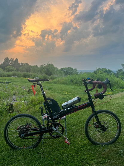 A black folding bike with a belt drive, red trim, and a red visibility triangle in the foreground of a picture with green hills and the clouds parting to reveal a fiery sunset in the middle. 