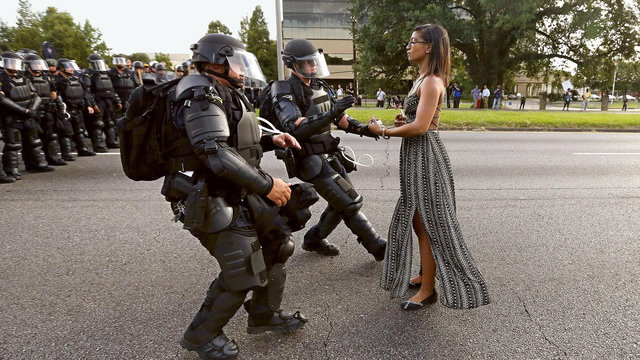 Black woman in a dress standing tall and poised facing the left while multiple cops in riot gear run forward to grab her by the arm to arrest her during Black Lives Matter protests