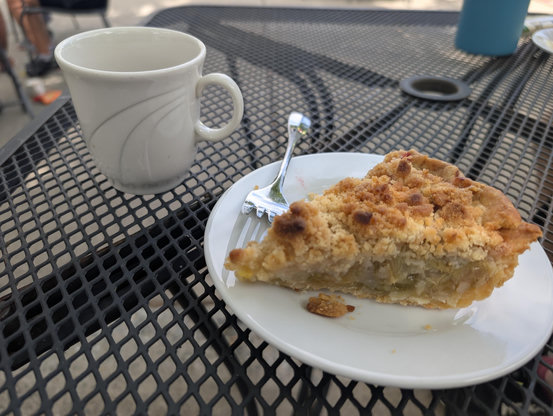 Slice of homemade pineapple and rhubarb pie on a plate with a fork.  Behind it a cup or coffee and a blue bottle.  All on a metal table.
