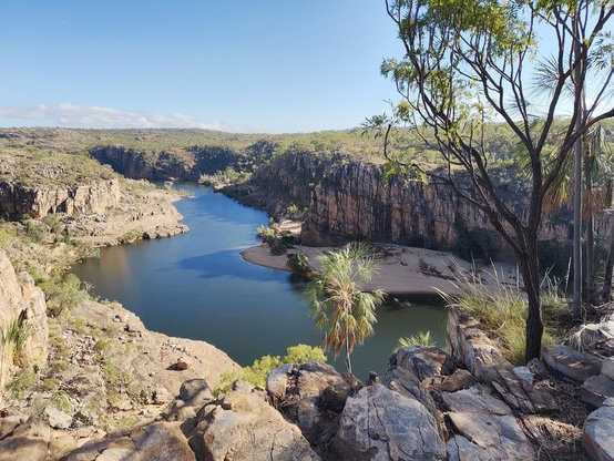 View down into Nitmiluk gorge from a rocky escarpment. Sky above is completely clear, the foreground harsh rocky boulders and way below, the river in a gorge