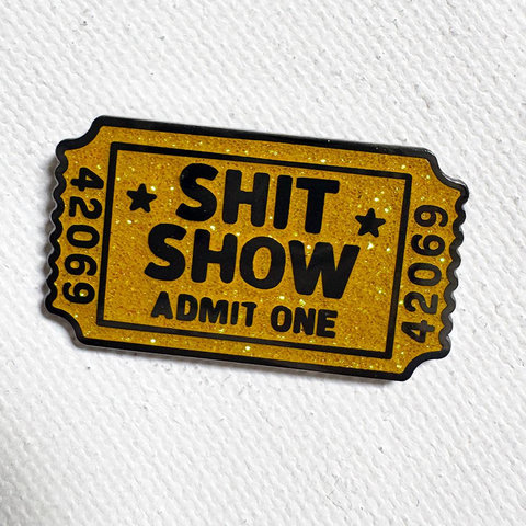 A glittery, rectangular gold ticket enamel pin that reads "SHIT SHOW" and "ADMIT ONE," with the number "42069" printed on both ends.