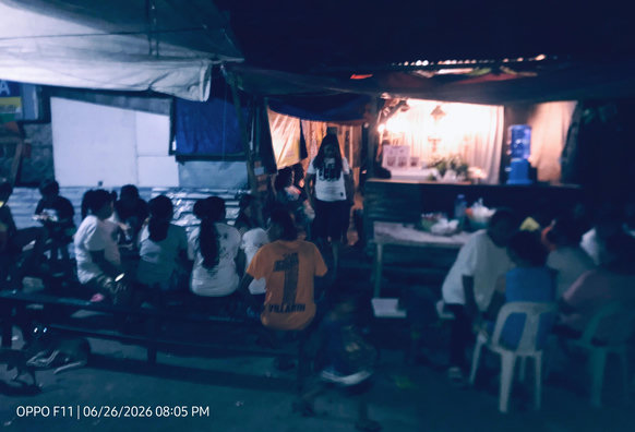 The scene depicts a community gathering in the Philippines, likely a funeral wake based on the attire and setup. Traditional Funeral wake are frequently held outdoors under tarps to accommodate many people.
A low-light photo taken at night shows a community gathering under a temporary tarp roof structure. 
A group of people sits on benches  On the right, other attendees sit on plastic chairs around a table. In the brightly lit background, a person stands near a white stage backdrop adorned with flowers.
 A timestamp at the bottom left reads "OPPO F11 | 06/26/2026 08:05 PM".