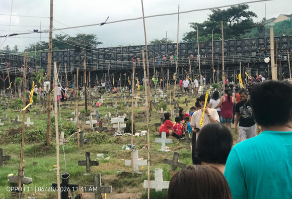 A public cemetery in the Philippines filled with numerous ground graves marked by simple wooden and concrete crosses, while a massive wall of multi-tiered columbarium niches looms in the background under a cloudy sky. People are scattered throughout the grassy graveyard (probably) visiting their departed loved ones, and thin bamboo poles with hanging wires crisscross above the scene.

