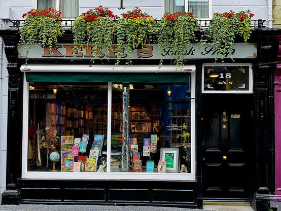 A photo of the front of Kerr’s Bookshop in Clonakilty with a beautiful display of flowers across the top and the address 18 Ashe Street painted on the glass above the front door. The shop front is glass with a book display and a glittery pillar. The awning above the window is retracted