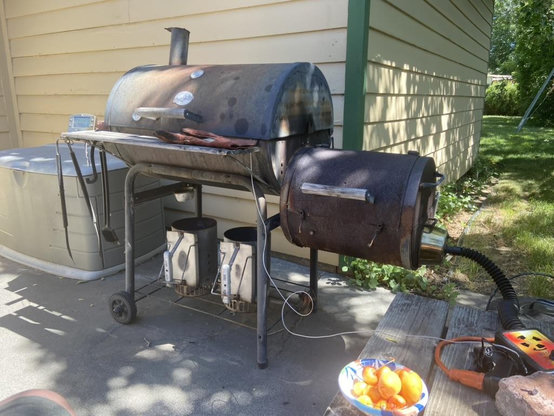 View of my patio.  An old (20 years?) CharGriller side-firebox grill/smoker with pork ribs cooking inside.  Two charcoal chimneys below. 
Lower right:  A bowl with locally grown Rainier cherries and a couple of apricots from my tree.  Also, the temperature-controlled blower used with the CharGriller.  Plum tree in the distant background.
Left:  The remote smoker thermometer can be seen.  The storage box with grill supplies at left.