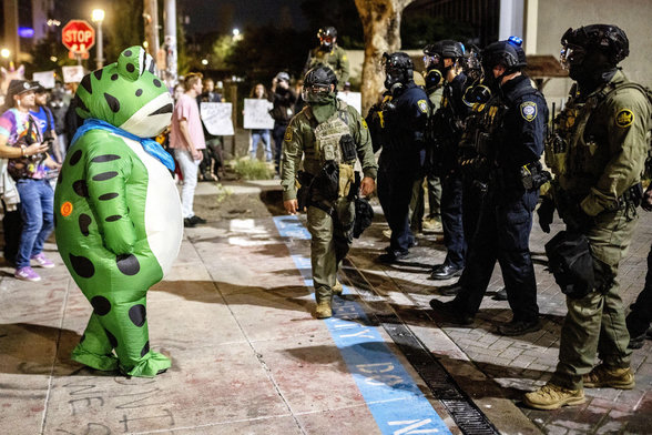 A person in a frog costume being confronted by armed police and soldiers at a No Kings rally.