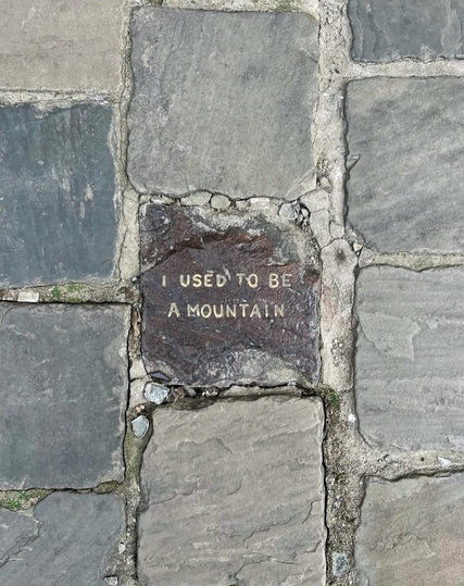 A top-down photograph of a cobblestone pavement. Centered among the grey and dusty stones is a single, darker, reddish-brown stone with carved or stamped text that reads in all-caps: "I USED TO BE A MOUNTAIN".
