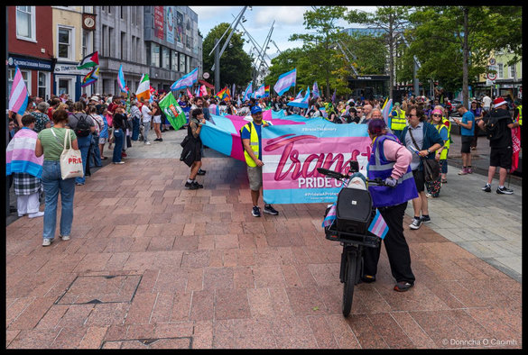 A cyclist with a speaker, marchers in a high-visibility vests holding a banner reading "Trans+ PRIDE CORK", "#BreakTheBinary", "#PrideIsAProtest" and "transpridecork.com" on a paved Cork street, with a crowd carrying trans flags behind and a "Cork PRIDE" tote bag on the left.
