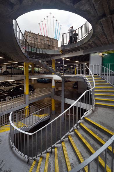 The Red Arrows fly over Edinburgh castle, trailing red, white, and blue smoke. We look up at it from a position half way down into a helical staircase that descends into Scotlands first multi-storey car park.
It’s an odd sort of composition, comprising a lot of yellow paint, a lot of concrete, some cars, a couple of people… but everything seems to be leading the eyes upwards to the sky above and the jets flying over us.
