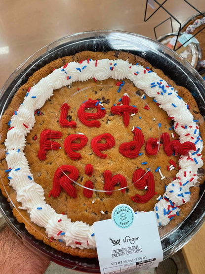 A giant cookie from Kroger inside a covered plastic tray. There’s a white frosting ring around the outside perimeter of the cookie, and inexpertly formed red frosting letters in the center which spell “Let Feedom (sic) Ring.” Red, white, and blue sprinkles are scattered around the cookie and icing, but most are strangely concentrated on the lower right - the 4:00-5:00 zone of the outer perimeter. 