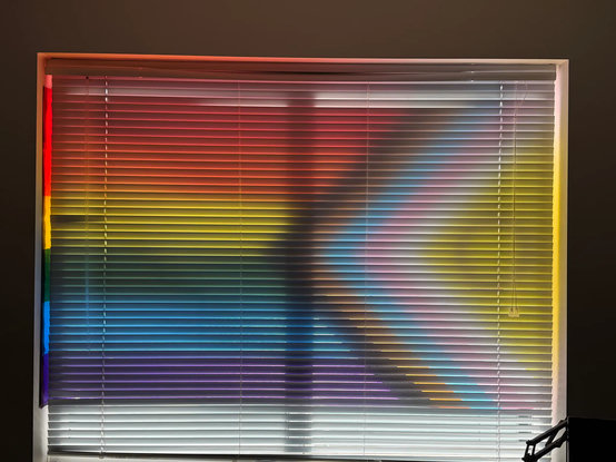 An apartment window with cheap metal blinds closed, showing off a very cool silhouette of the progress pride flag put up across both windows, in front of the blinds.
