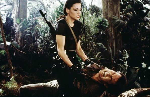 photo from Goldeney of Xenia Onatopp (white woman with dark hair in a black t-shirt and black pants with a gun on her back) straddling James Bond (Pierce Brosnan) in a jungle as he screams in pain