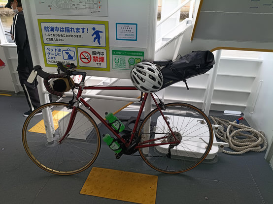 A bicycle in the cargo area of a tiny ferry, with various notices in Japanese on the wall behind.