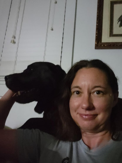 A 100 lb black labrador retreiver is laying behind a woman while panting in fear. She is reaching up to stroke him and comfort him.