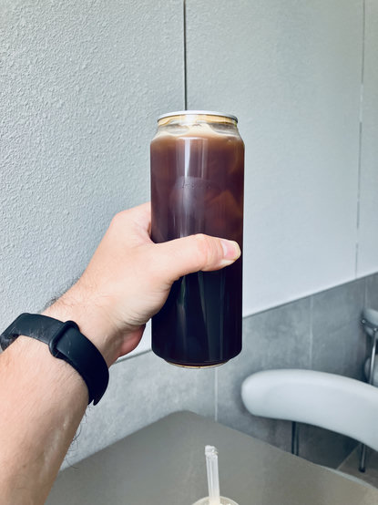 A hand wearing a black fitness band holds a tall, clear can filled with a dark iced beverage, with several ice cubes visible near the top. The photo appears to be taken inside a modern café or restaurant, with a gray textured wall, gray tabletop, and a white chair softly blurred in the background. A wrapped straw rests on the table below the drink.