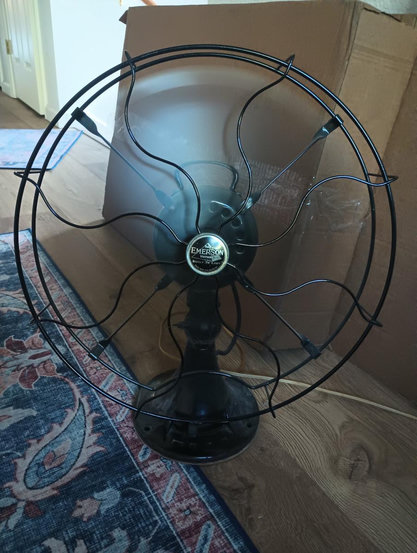 A color portrait photo of an old metal floor fan. It dates to around the 1920s, likely. It is black and has a small medallion in the center. The fan is running as the blades are just a blur. A cardboard box is seen in the background.