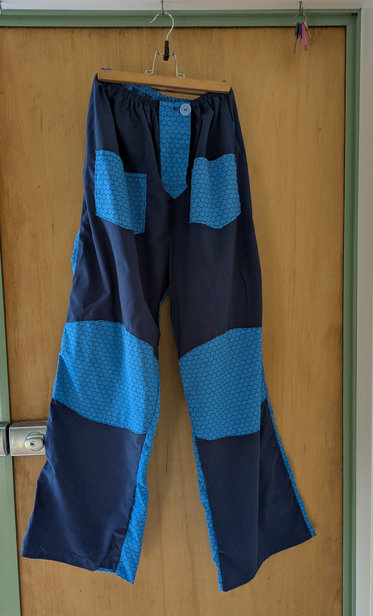 A pair of wide-legged trousers on a hanger. The front of the trousers is dark blue. It has pockets and knee patches in a lighter blue colour with a black honeycomb pattern. The back of the trousers is entirely made of the honeycomb fabric. The background is a wooden door with a pale green frame.