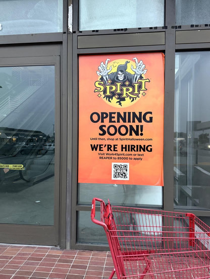 Orange Spirit Halloween "Opening Soon" sign on storefront window with grim reaper logo and hiring info. Red shopping cart in foreground.