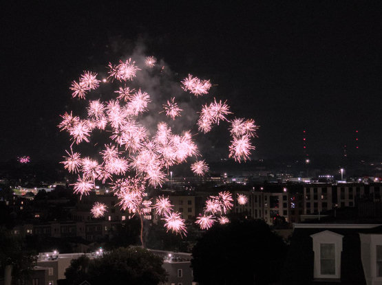 A sparkling fireworks burst viewed from Telegraph Hill in Boston. The trail of the initial rocket launch is still faintly visible, showing the rocket's trajectory. Dorchester is visible in the background over South Boston rooftops, and there is another burst going off in the far distance.