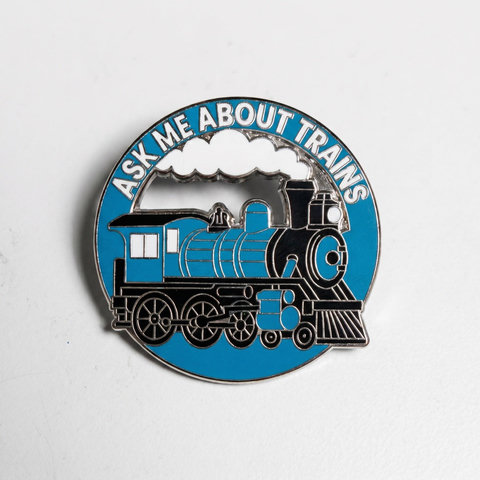 A circular enamel pin featuring a blue steam locomotive with a white cloud of smoke and the text "ASK ME ABOUT TRAINS" arched across the top.