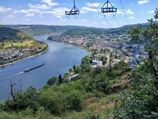 La vallée du Rhin à hauteur de Boppard (à droite) de la photo photographiée depuis le sentier de randonnée Saar-Hunsrück-Steig qui se transforme en sentier approximatif à travers le massif schisteux. Ce sentier fait partie d'un sentier d'escalade. Il suit sur 700 mètres le tracé du télésiège. Une péniche navigue sur le Rhin, un autre village est visible sur l'autre rive à gauche. On peut voir plusieurs sièges du télésièges qui montent et descendent en permanence tout au long de la journée. On peut aussi voir les voies de chemins de fer de la ligne du Rhin (rive gauche). 

The Rhine valley near Boppard (right) in the photo photographed from the Saar-Hunsrück-Steig hiking trail which turns into an approximate trail through the shale massif. This trail is part of a climbing trail. It follows the route of the chairlift for 700 meters. A barge sails on the Rhine, another village is visible on the other bank on the left. Several chairlift seats can be seen constantly moving up and down throughout the day. You can also see the railway tracks of the Rhine line (left bank).