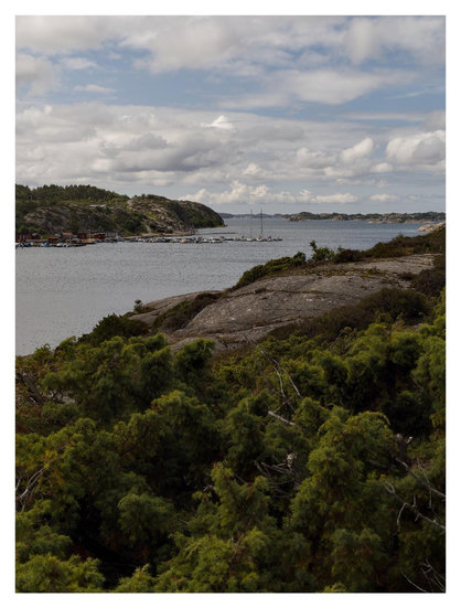 En stÄende bild pÄ havet mellan uddar och öar. En bit ut ligger en brygga med fritidsbÄtar. Himmelen Àr molniga med blÄ inslag. I förgrunden ett buskage före klippor mot havet.