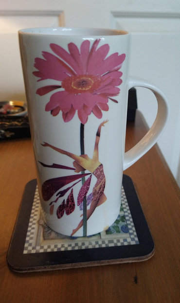 Tall mug of tea on a bedside with a pink gerbera flower in the centre and a leaping faerie below