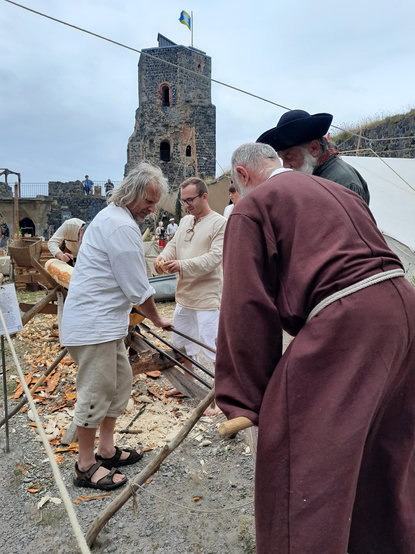 In einem Burghof, Personen in historischen Kostümen arbeiten an einem Baumstamm, im Hintergrund ein Burgturm.