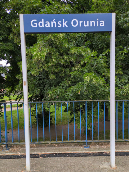 Tabliczka peronowa "Gdańsk Orunia". W tle drzewa za niebieskim płotem.