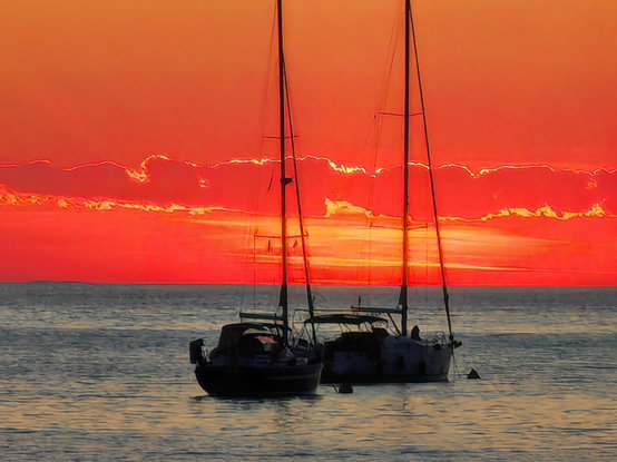 A pair of sailing yachts moored offshore. The sea is calm and the two yachts are almost silhouetted against the sky which is a brilliant orange red colour as the sun sets.