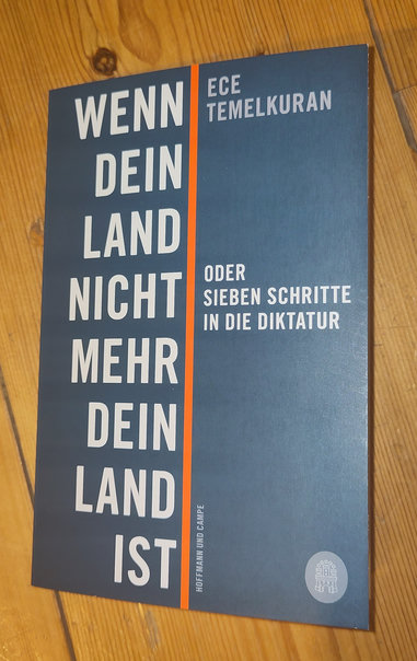 Grünes Buch, weiße Schrift links neben mittigem roten, dünnen Balken:
Wenn dein Land nicht mehr dein Land ist"
Rechts vom Balken: "Ece Temelkuran"
darunter: "Oder sieben Schritte in die Diktatur"
Das ganze auf Dielenboden liegend. 