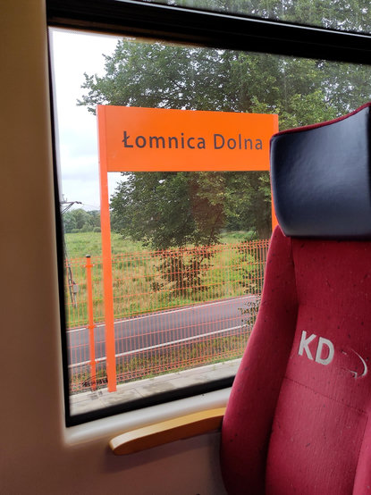Widok przez okno pociągu Kolei Dolnośląskich, co można poznać na napisie na fotelu. Za oknem tabliczka z nazwą stacji Łomnica Dolna. Zarówno tablica jak i płot za nią są koloru jaskrawo pomarańczowego.
