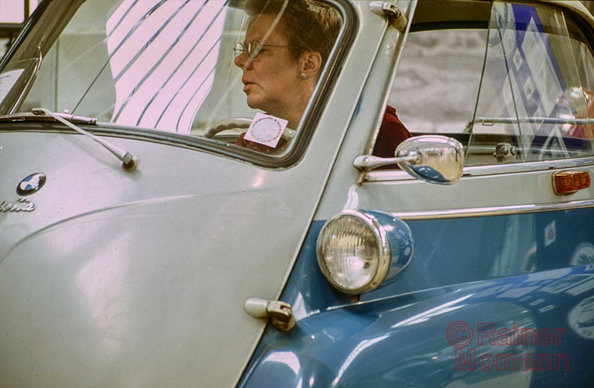 Farbfotografie im Format 3:2 Landscape.
In einer Sonderausstellung des Deutschen Museums. Abgebildet ist der größte Teil einer BMW Isetta 250. In der linken Bildhälfte die kugelige Front. Rechts die linke Seite des Fahrzeugs mit der klassischen blauen Farbe unterhalb des Schiebefensters. Im Inneren der Isetta sitzt eine Person.