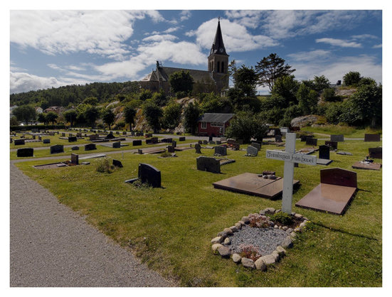 En liggande bild pÄ en kyrkogÄrd med gravstenar. PÄ berget bredvid ligger kyrkan. Himmelen Àr blÄ med vita moln.