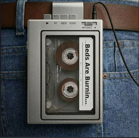 A brushed-silver, vintage-style portable cassette player clipped to a person's brown leather belt worn with blue denim jeans. A clear rectangular window on the front reveals a cassette tape inside, which features a white label with the text "Beds Are Burnin..." printed vertically, along with "TYPE I NORMAL POSITION". The top edge of the player features mechanical buttons for Play, Fast Forward (FF), Rewind (REW), and Stop, as well as a function slider. A black audio cable is plugged into a jack on the top right corner and drapes down the side.
