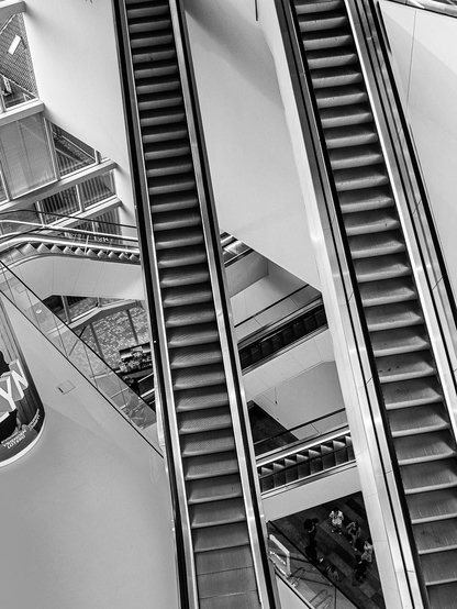 Black-and-white photograph looking down at a steep pair of parallel escalators inside a multi-storey building atrium, framed diagonally between angled white walls and glass balustrades. Additional escalators and railings are visible on lower levels below, with several small figures walking on a patterned floor at the bottom. A partial illuminated sign with large letters is visible in the lower left corner, and glass-walled office floors are visible in the upper left background.