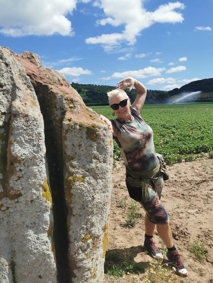Some crazy woman in crew cut and dark glasses poses beside a bronze age standing stone in the middle of a crop field