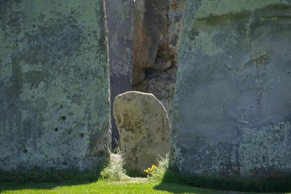 Weathered stone standing between large, worn stones, with grass and small yellow flowers at its base.
